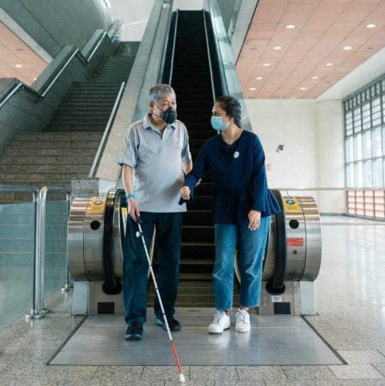 a visually-impaired commuter being assisted by a Caring Commuter Champion at the escalator of an MRT station